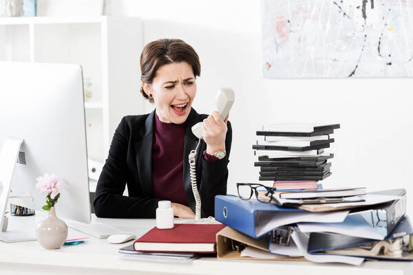 angry attractive businesswoman screaming while talking by stationary telephone in office