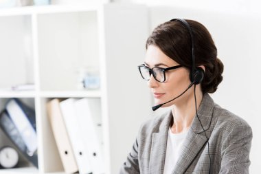 attractive call center operator in glasses and headset looking away in office clipart