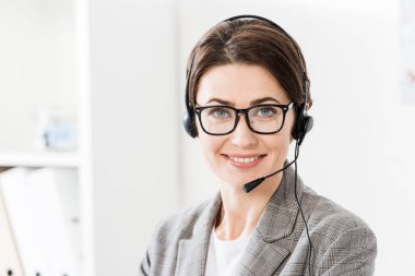 smiling beautiful call center operator in glasses and headset looking at camera in office clipart