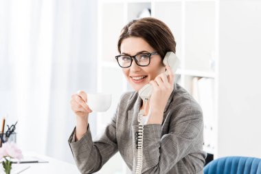 Sabit telefonla konuşurken ve ofiste kahve içme gülümseyen çekici iş kadını