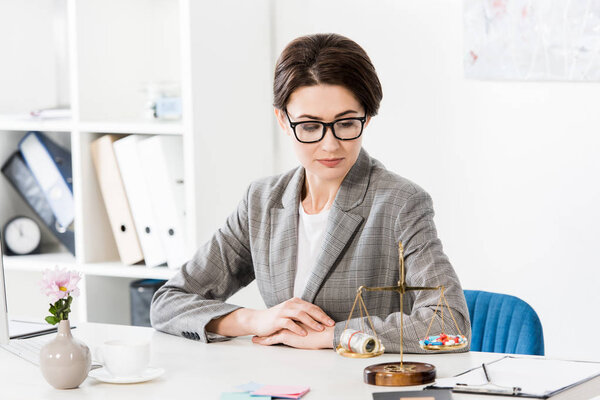 attractive lawyer looking at justice scales with money and drugs on table in office