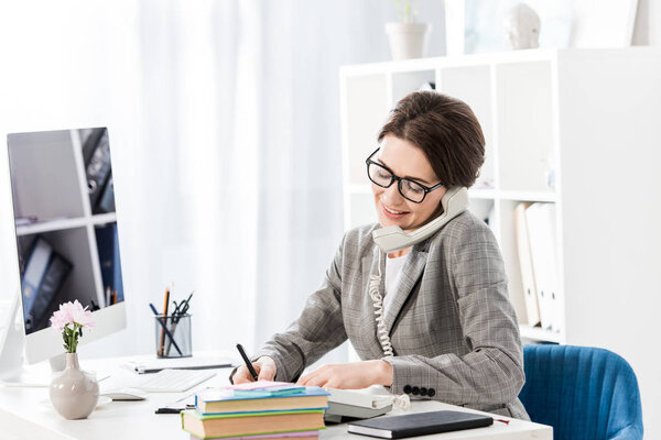 smiling attractive businesswoman talking by stationary telephone in office