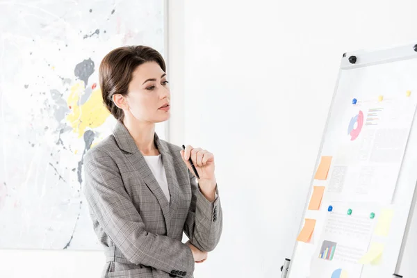 オフィスのプロジェクトのプレゼンテーション中にフリップチャートを見て灰色のスーツの魅力的な女性実業家物思いにふける — ストック写真