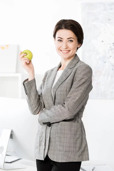 Sonriente Atractiva Empresaria Traje Gris Sosteniendo Manzana Mirando Cámara Oficina —  Fotos de Stock