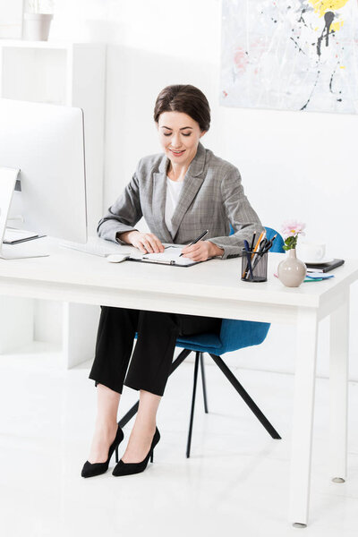attractive businesswoman in grey suit writing something to clipboard at table in office