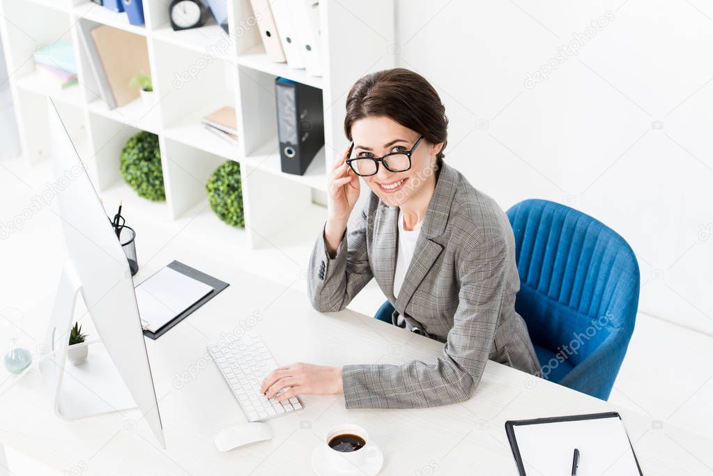 high angle view of smiling attractive businesswoman using computer at table in office and looking at camera