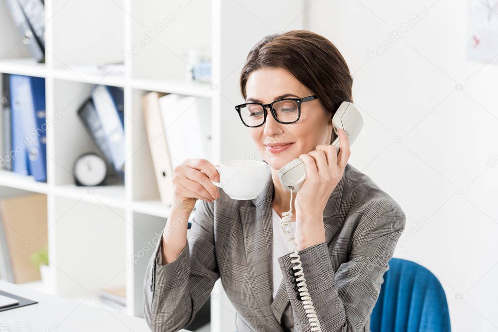 smiling beautiful businesswoman talking by stationary telephone and drinking coffee in office