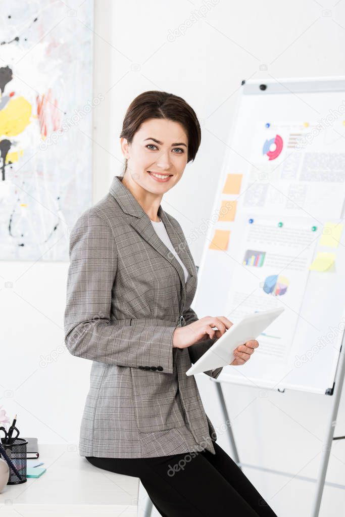 smiling attractive businesswoman in grey suit sitting on table and using tablet in office