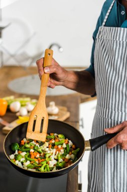 cropped image of middle aged man cooking vegetables on frying pan in kitchen clipart