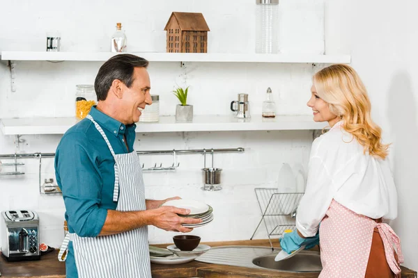 Mature Wife Washing Dishes Husband Holding Plates Kitchen — Stock Photo, Image