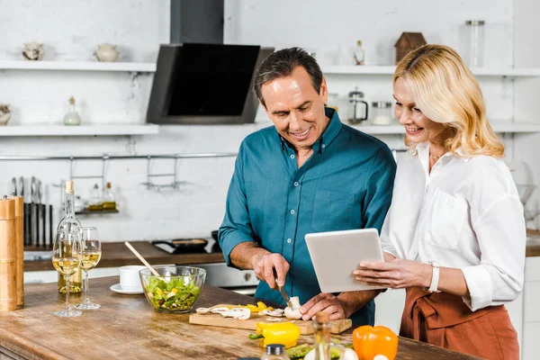 成熟的妻子使用平板电脑和丈夫削减蔬菜在厨房 — 图库照片