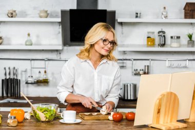 çekici Olgun kadın mutfakta kesme mantar tarifi kitabı bakarak