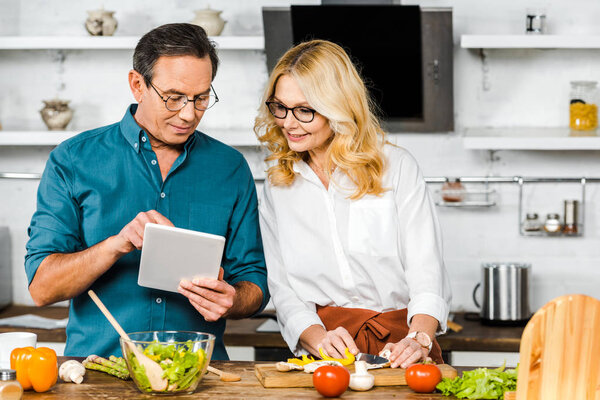 mature husband showing something on tablet to wife in kitchen