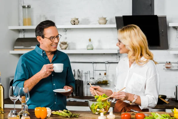 成熟した妻はサラダを混合と夫のキッチンでお茶を飲みながら笑みを浮かべて — ストック写真