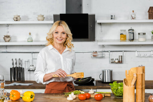 attractive middle aged woman holding frying pan for cooking vegetables in kitchen, looking at camera