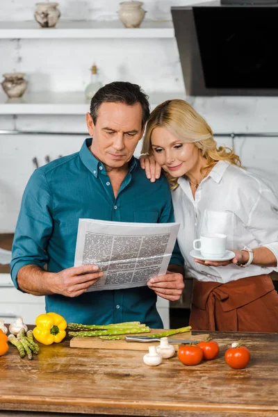 成熟的妻子拿着一杯茶和阅读报纸一起在厨房里 — 图库照片