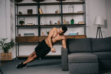 mixed race man doing plank exercise in living room near sofa clipart