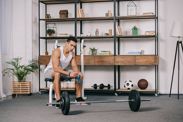 pensive bi-racial man sitting near barbell and holding sport bottle 