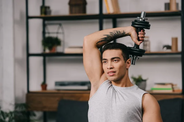 bi-racial man with tattoo in hand holding dumbbell over head