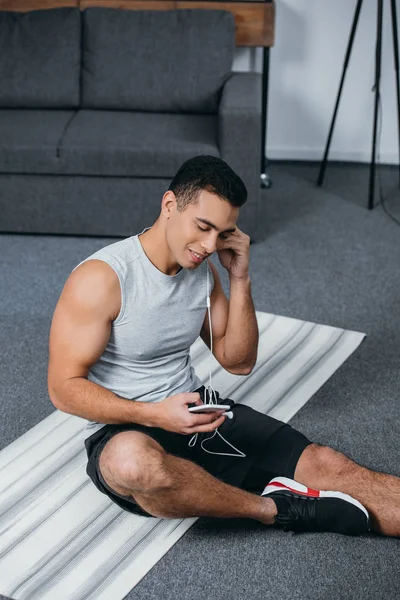 happy bi-racial man putting earphones in ear and holding smartphone while sitting on  fitness mat