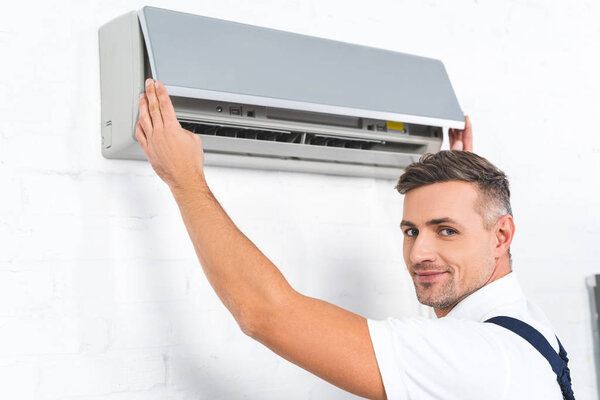 handsome repairman checking air conditioner and smiling at camera 