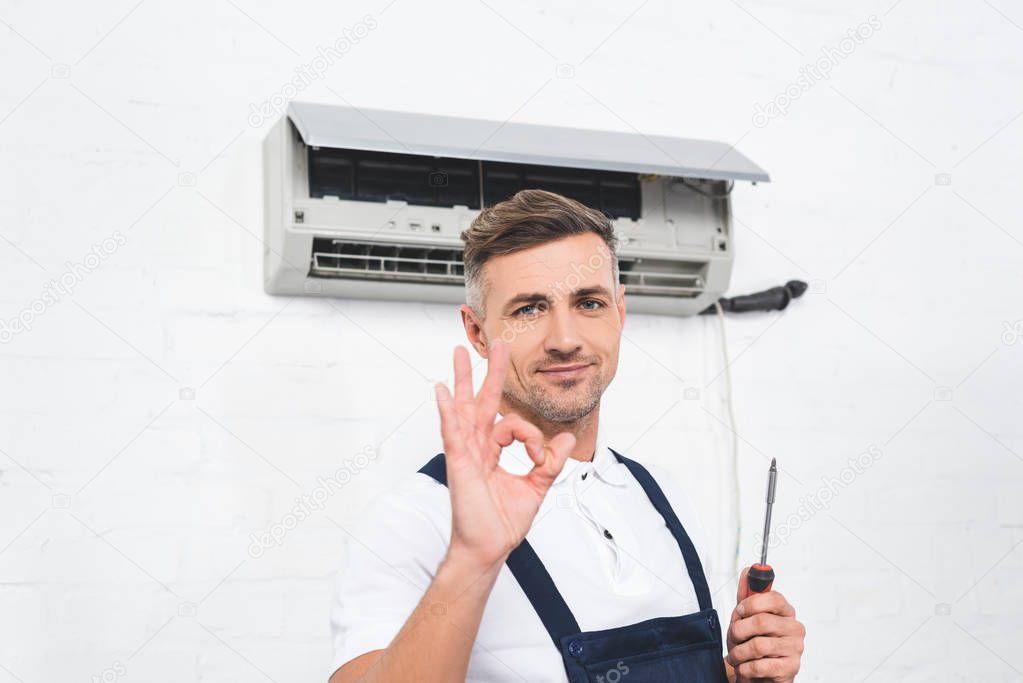 adult repairman holding in hand screwdriver and showing okay gesture near air conditioner 