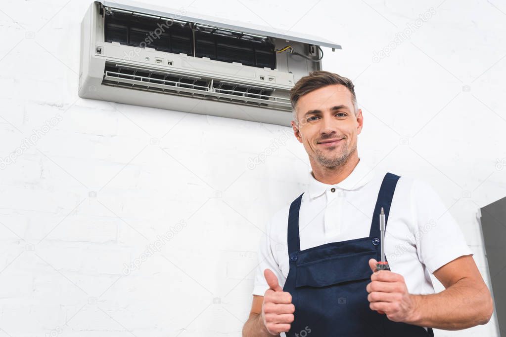 Handsome repairman showing thumb up gesture with air conditioner at background