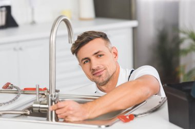 smiling adult repairman with spanner repairing faucet at kitchen and looking at camera clipart