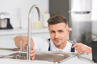 smiling adult repairman holding pipe and spanner while repairing faucet at kitchen and looking at camera clipart