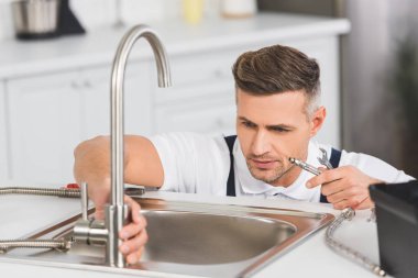 adult repairman holding pipe and spanner while repairing faucet at kitchen clipart