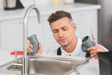 thoughtful adult repairman holding pipes and tools while repairing faucet at kitchen  clipart