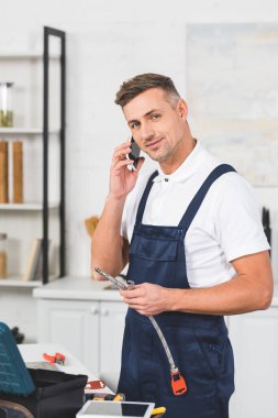Yetişkin tamirci holding boru tamir ve smartphone üzerinde konuşurken kameraya bakarak mutfak musluk için gülümseyen