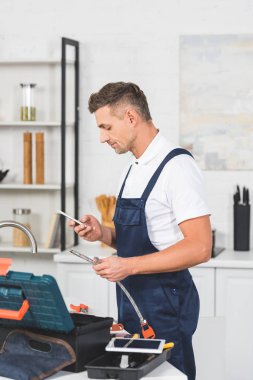 Profil sahibi yetişkin tamirci boru tamir ve smartphone kullanarak mutfak musluk için holding
