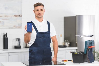 smiling adult repairman showing smartphone with Facebook app while standing at kitchen and looking at camera 