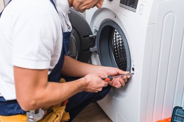 cropped view of adult repairman working with screwdriver while repairing washing machine in bathroom  clipart