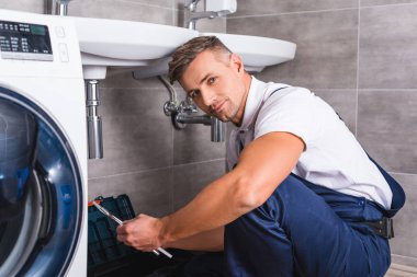 katta oturan ve onarım aracı, banyo ve kameraya bakarak tutan yetişkin tamirci
