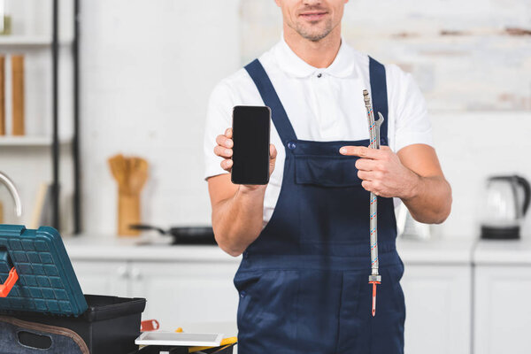 cropped view of smiling adult man holding pipe and spanner while pointing at smartphone wit blank screen
