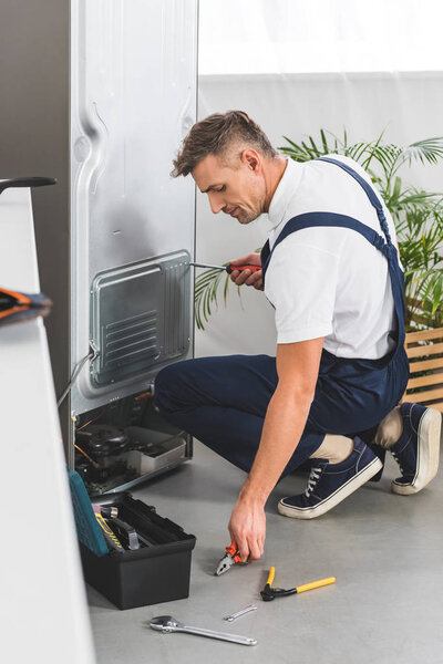 взрослый мужчина ремонтирует холодильник на кухне и берет плоскогубцы с пола
