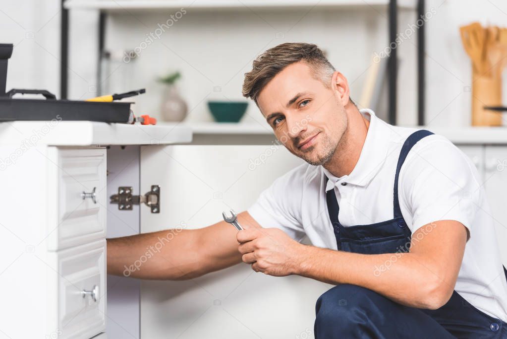 adult repairman repairing pipes of sink by spanner at kitchen and looking at camera