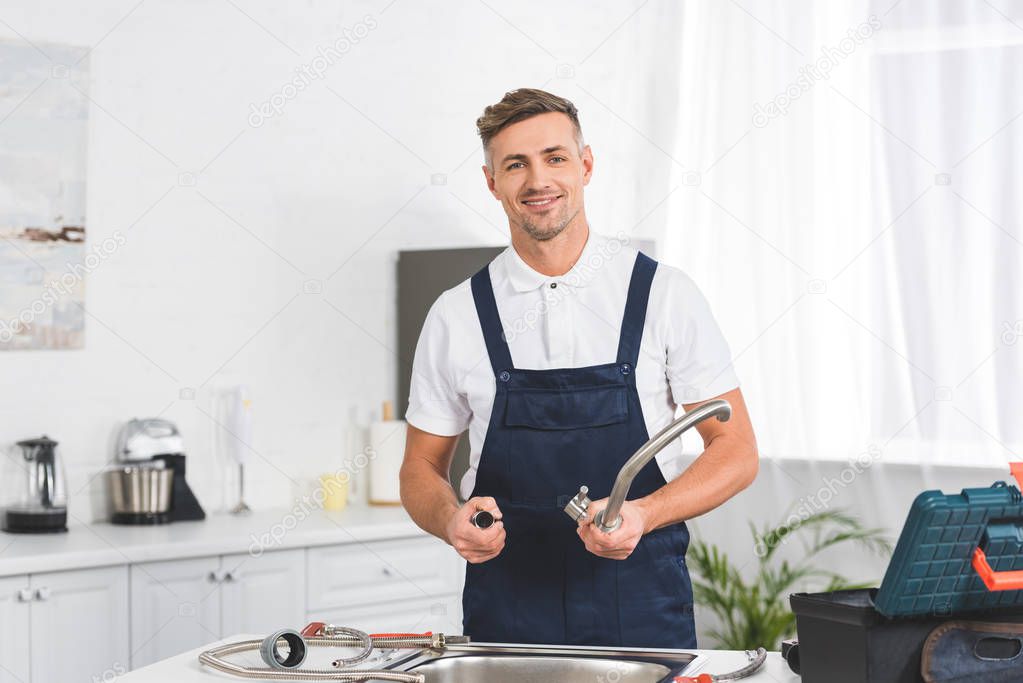 smiling adult repairman taking off kitchen faucet for repairing and looking at camera 