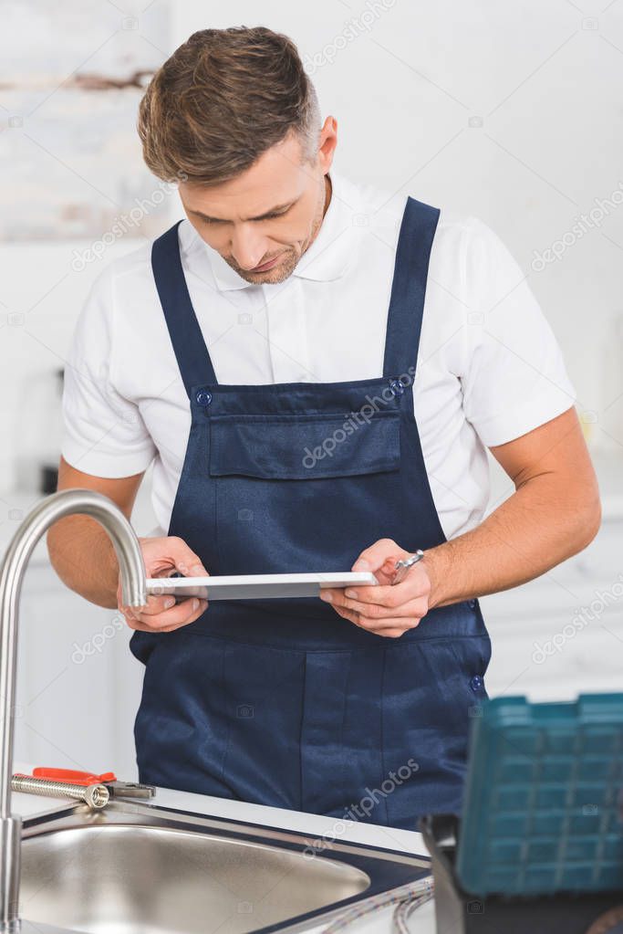 thoughtful adult repairman holding spanner and looking at digital tablet while repairing kitchen faucet   