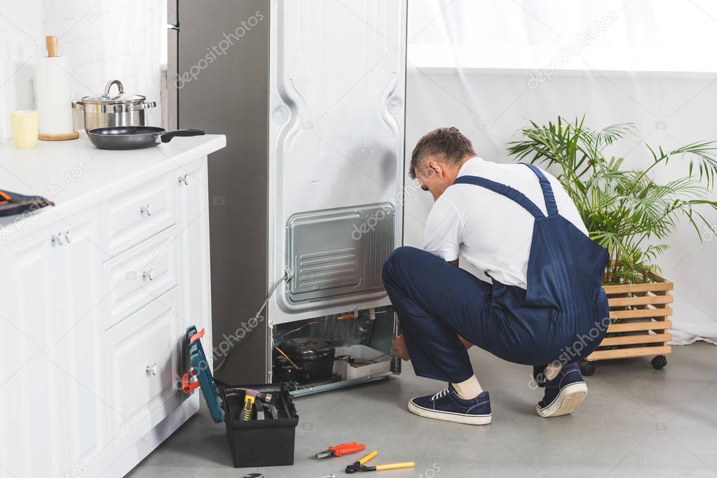 adult handyman repairing refrigerator at kitchen with tools on floor