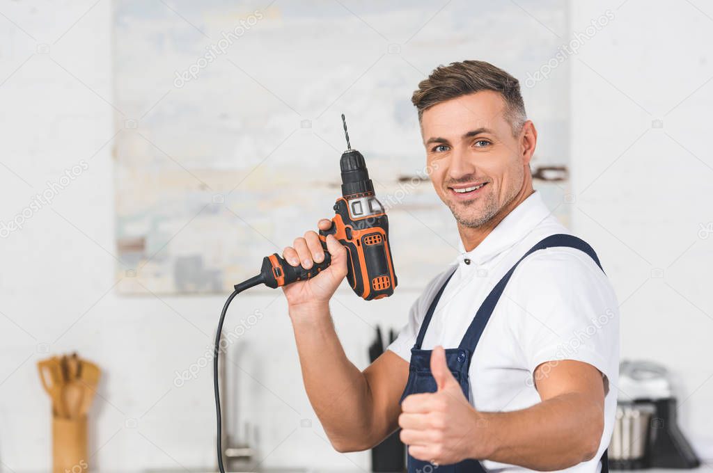 selective focus of smiling adult repairman holding drill in hand and showing thumb up sign 