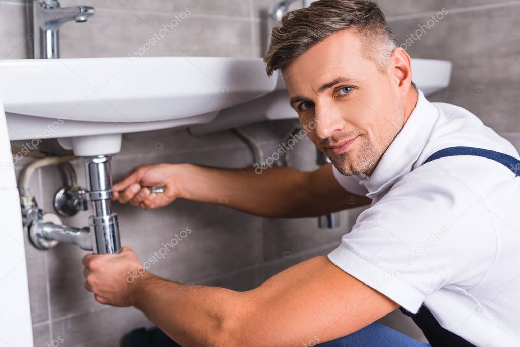 adult repairman repairing sink at bathroom and looking at camera