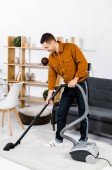 schöner Mann im modernen Wohnzimmer lächelt und putzt Haus mit Staubsauger 