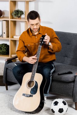handsome man in glasses sitting on sofa and tuning acoustic guitar at home clipart