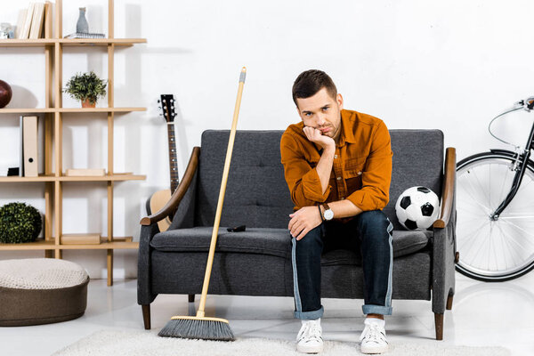 sad man sitting on sofa with broom and ball in modern living room 
