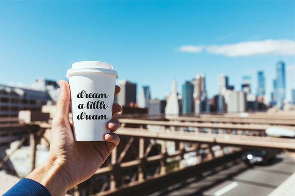 partial view of man holding disposable cup of coffee with "dream a little dream" lettering and blurred new york city on background