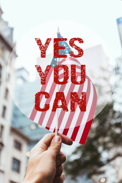 cropped shot of man holding american flag in hand with blurred new york city street on background and "yes you can" lettering