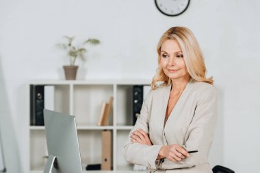 çapraz kolları ile ayakta ve Office masaüstü bilgisayar bakarak güzel olgun iş kadını